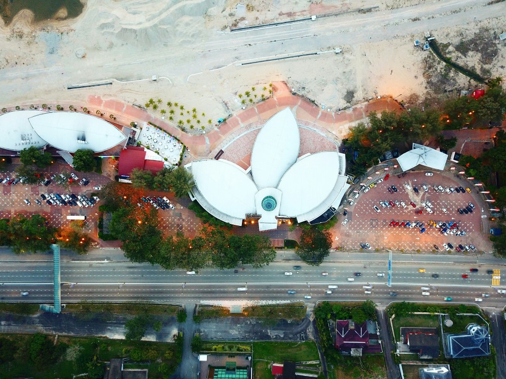 Aerial view of a lotus-shaped building in Johor Bahru, Malaysia, surrounded by parking lots and sandy construction areas near the Singapore border.