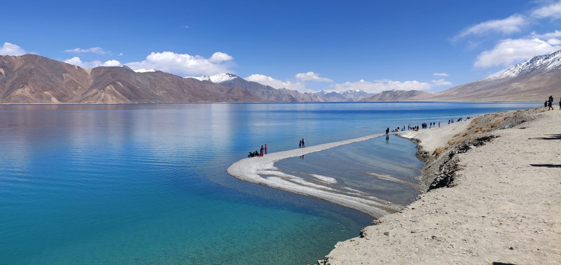 Tourists walking along the pristine shoreline of Pangong Lake in Ladakh, surrounded by turquoise waters and snow-capped Himalayan mountains under a clear blue sky.