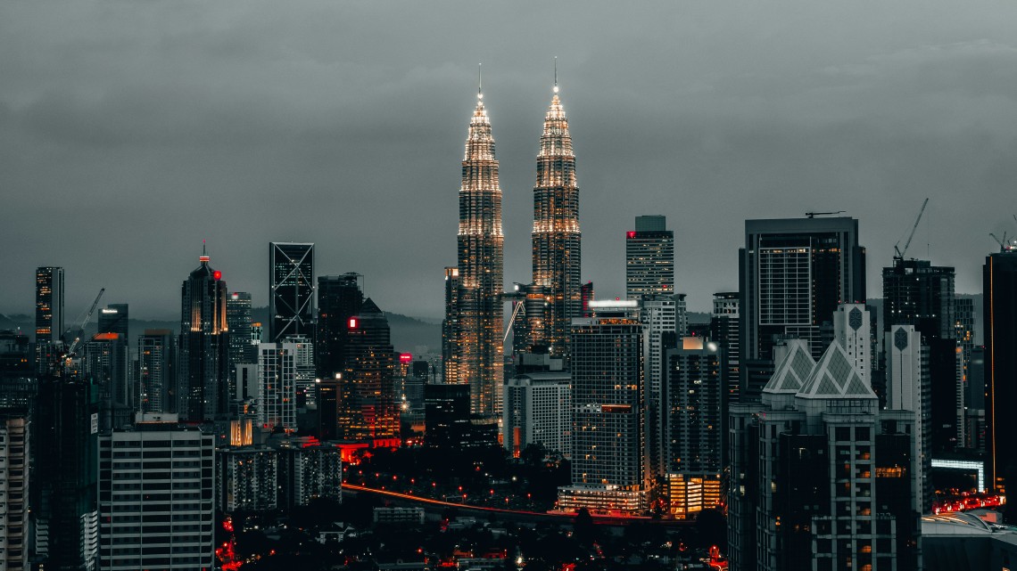 Nighttime view of Kuala Lumpur city skyline featuring the illuminated Petronas Twin Towers under a cloudy sky.
