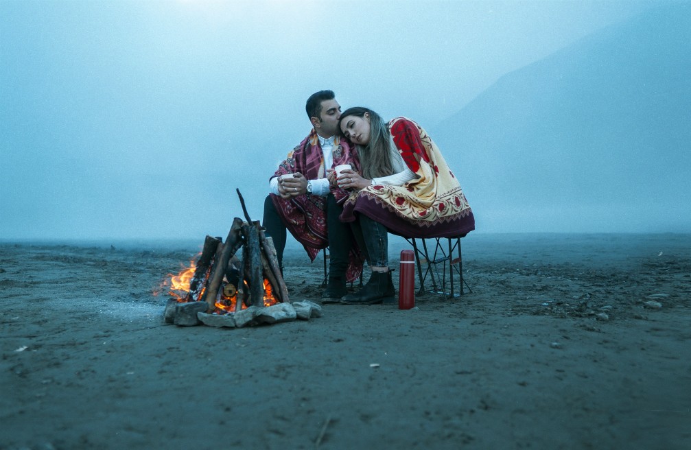 Couple warming up by a fire after a long day of trekking, with rugged terrain all around.