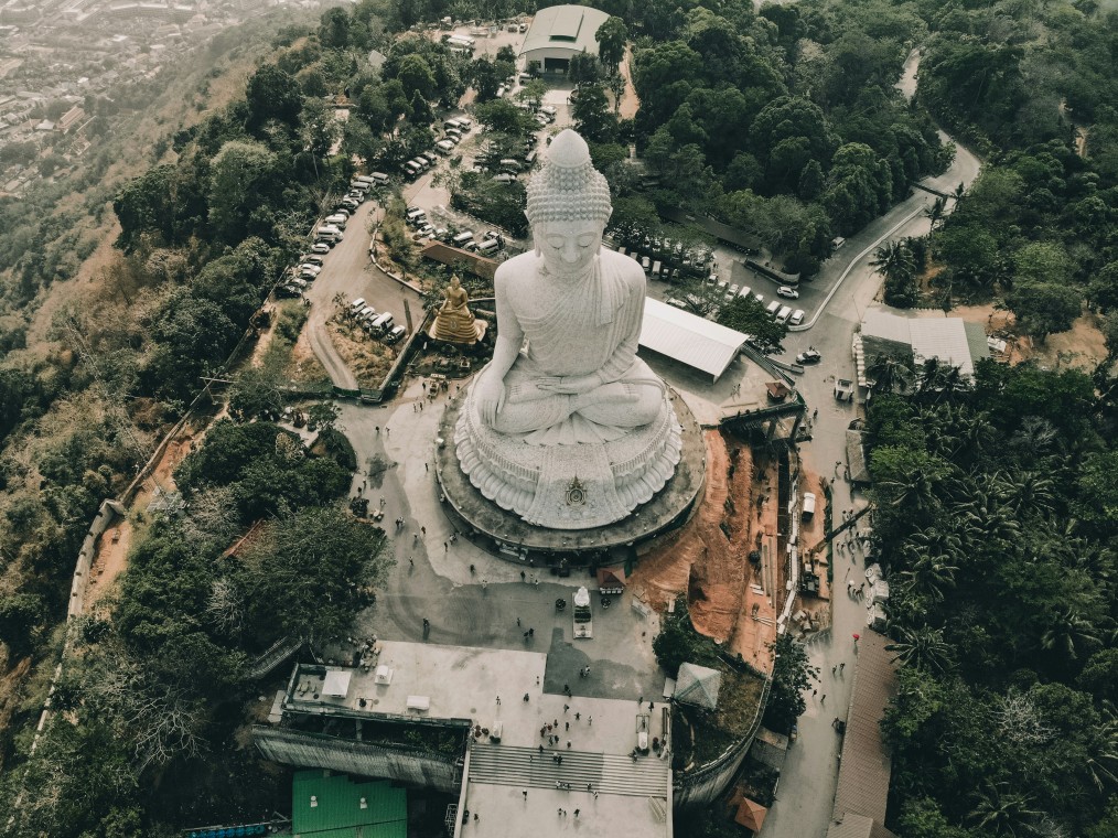 Aerial view of the iconic Big Buddha statue in Phuket, Thailand, surrounded by lush greenery and hilltop roads, with visitors exploring the site below.