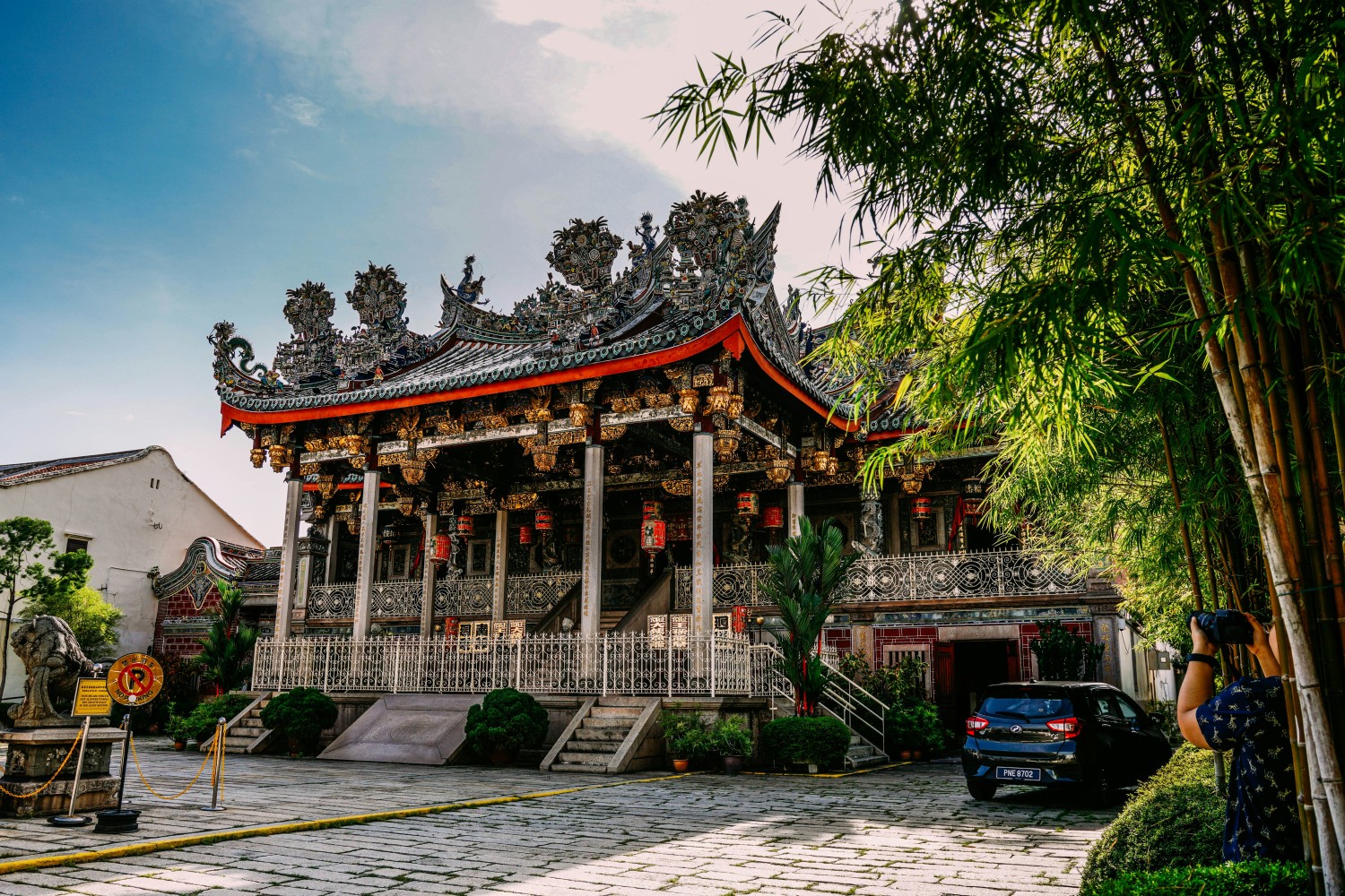 Traditional Chinese temple with ornate rooftop carvings and red lanterns in George Town, Penang, surrounded by lush bamboo and greenery.