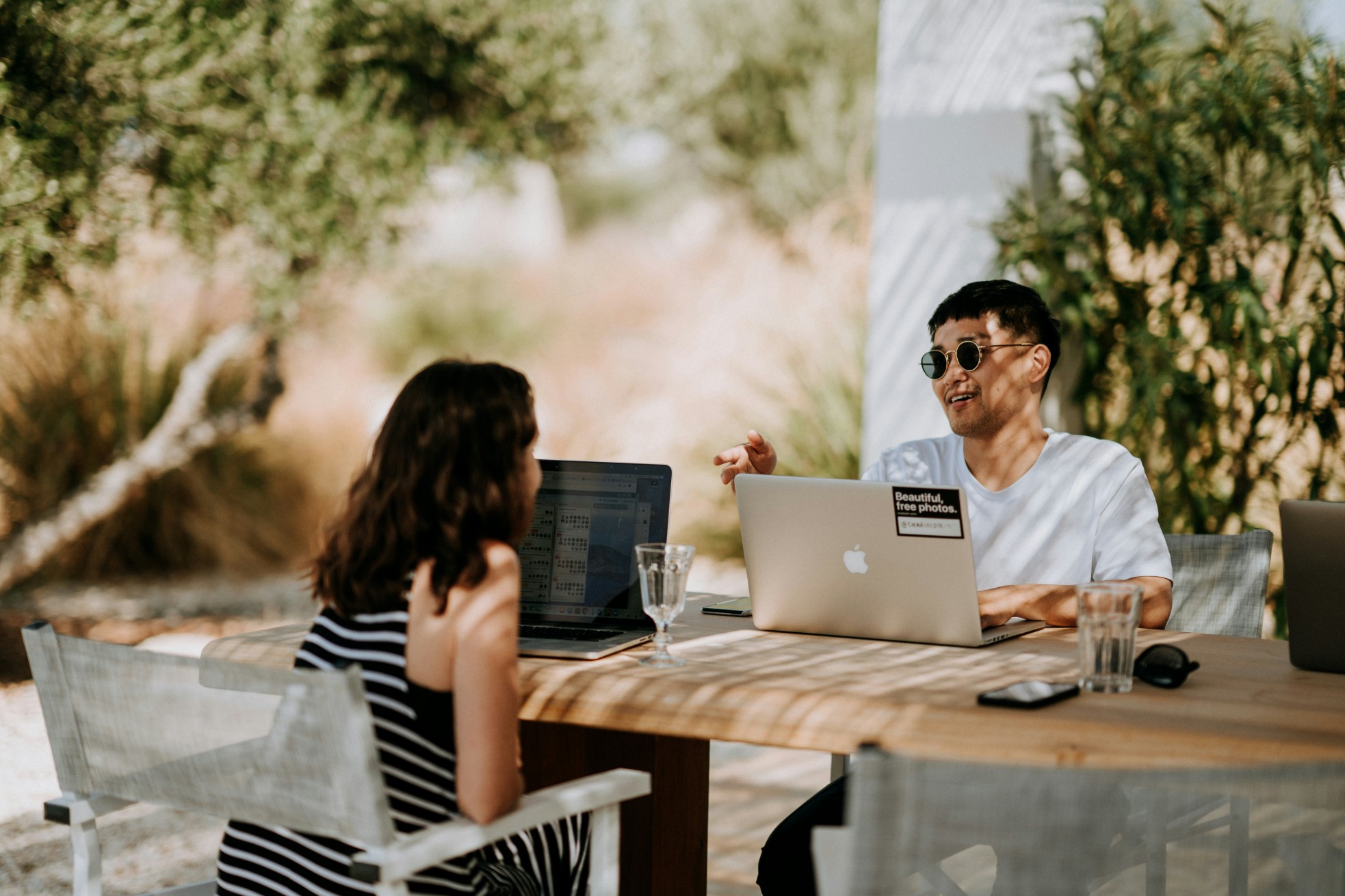 Two people enjoying a workation, working on laptops and talking at an outdoor wooden table in a sunny setting.