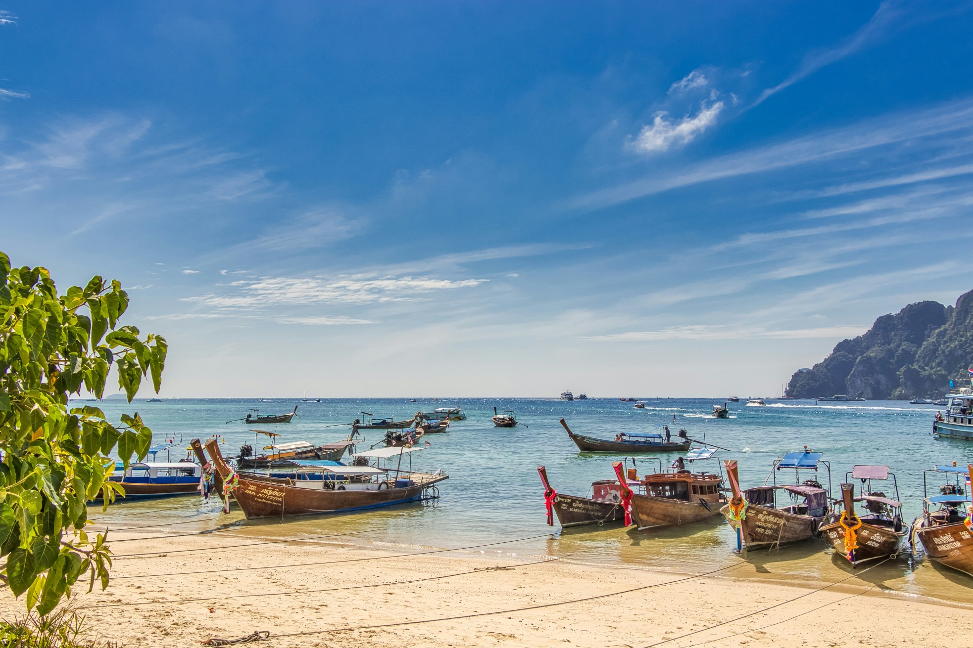 Traditional long-tail boats anchored along the sandy beach in Krabi, Thailand, with clear blue waters and limestone cliffs in the distance.