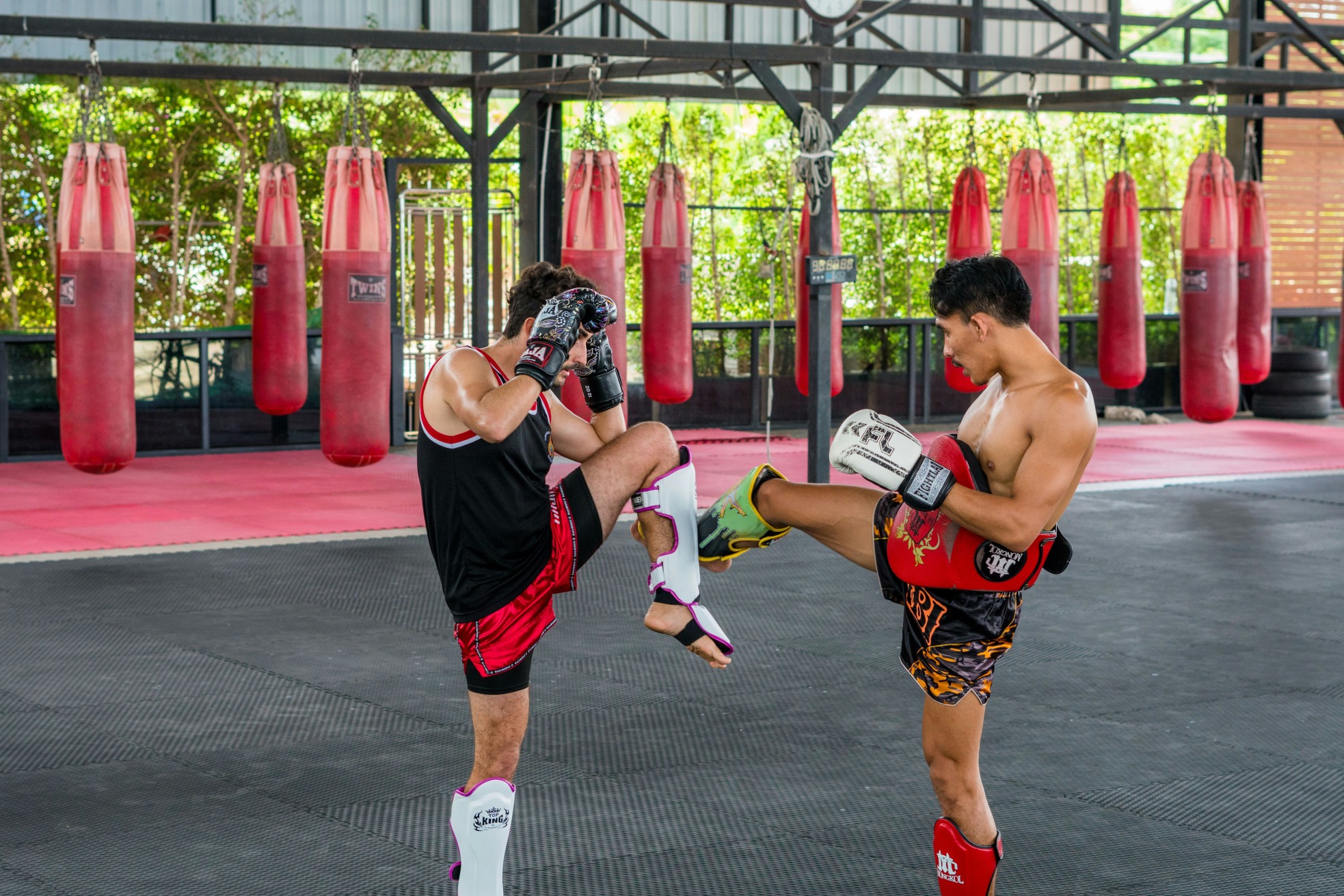 Two men practicing Muay Thai sparring in a gym with red punching bags in the background.