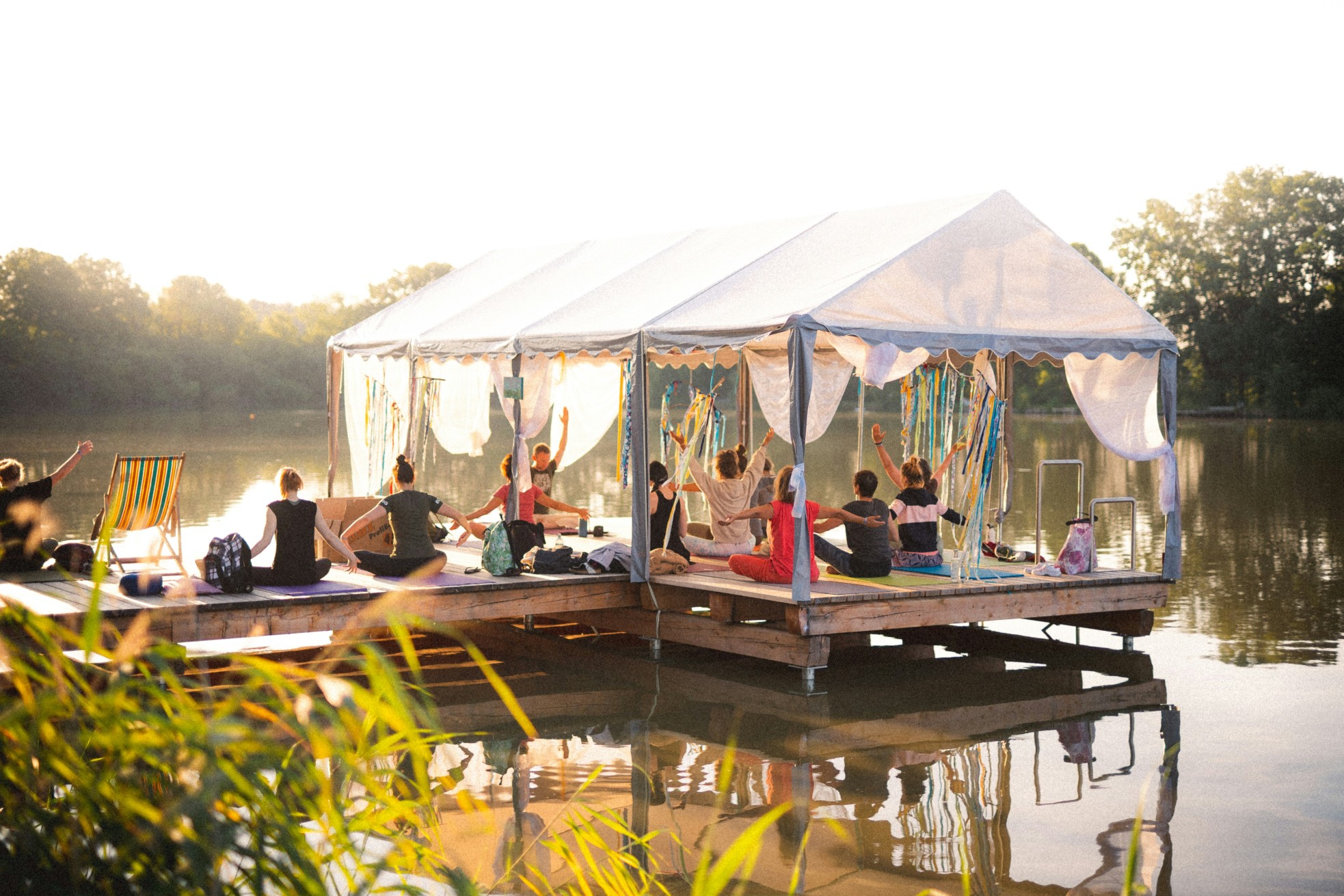 Group of people doing yoga under a canopy on a floating dock by the lake at sunset.