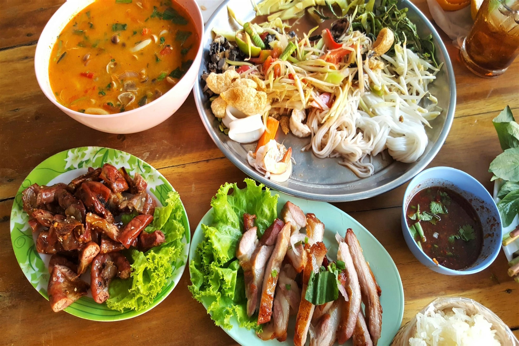 Thai food spread with spicy soup, grilled meat, noodles, fresh vegetables, and dipping sauce on a wooden table.